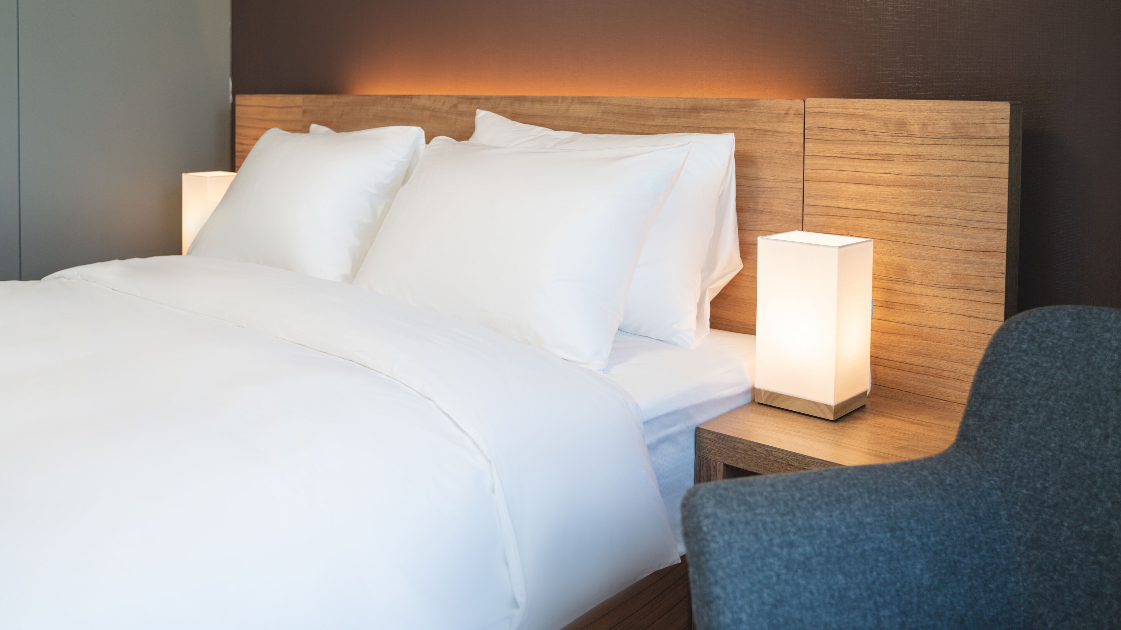 A neatly made bed with white linens and pillows, a bedside table with a lamp, and a blue chair.