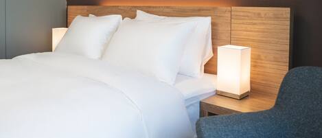 A neatly made bed with white linens and pillows, a bedside table with a lamp, and a blue chair.