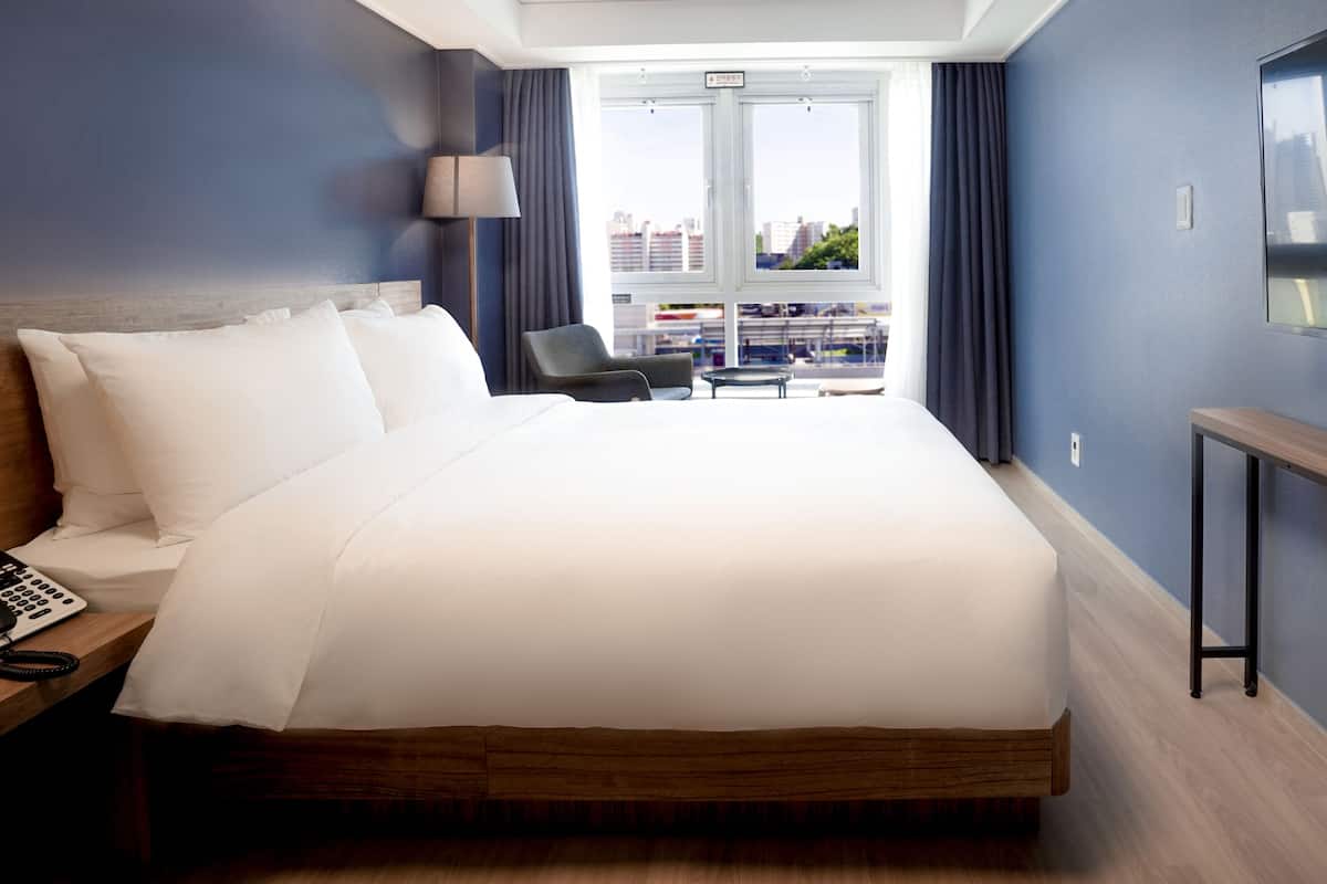 Habitación de hotel con una cama grande, un escritorio con teléfono, televisión en la pared y vistas a edificios a través de la ventana.