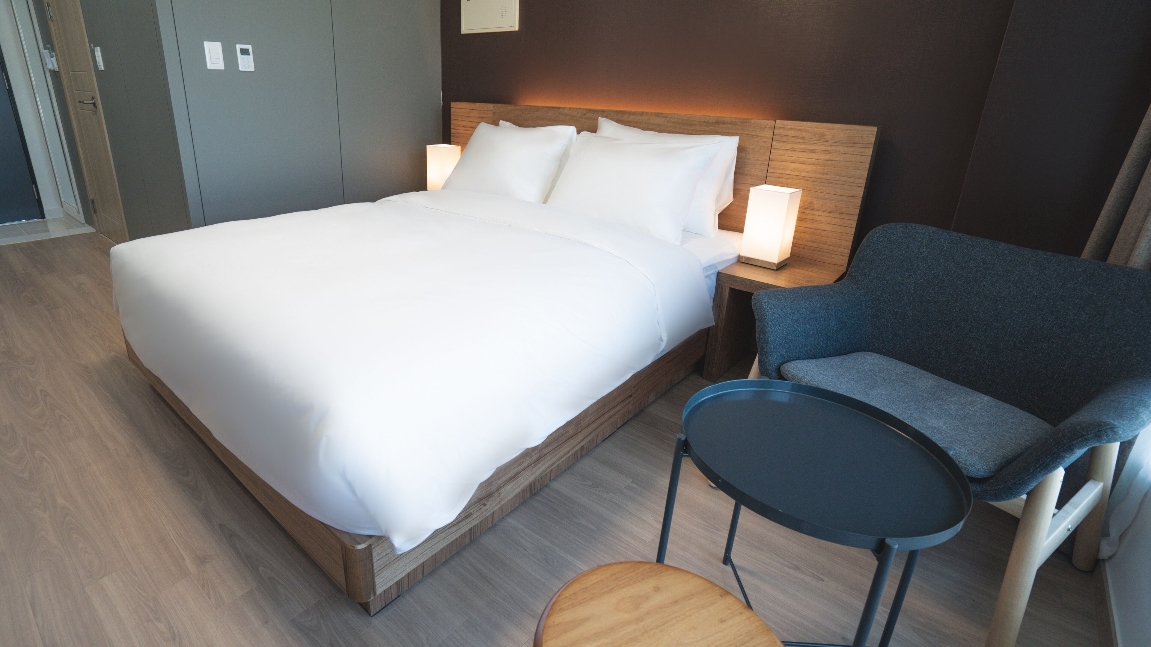 Quarto de hotel com uma cama grande, uma cadeira, uma mesinha e uma mesa de cabeceira com abajur.