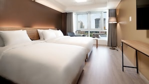 Een hotelkamer met twee bedden, een klein tafeltje, een lamp en uitzicht op de stad door grote ramen.