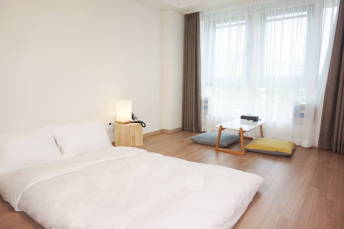 Una habitación de hotel con una cama grande, una mesita de noche con lámpara, una pequeña mesa con una silla y vistas al exterior a través de cortinas translúcidas.