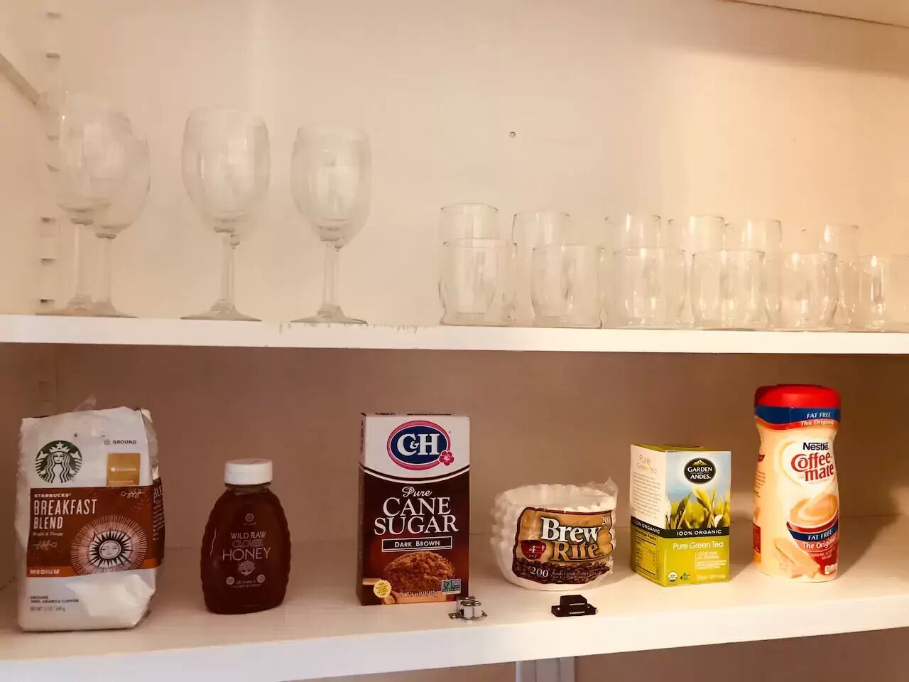 Una estantería con diversos objetos, incluyendo una bolsa de Starbucks, una botella de miel, una caja de azúcar de caña, un tarro de café y varios vasos de vino.