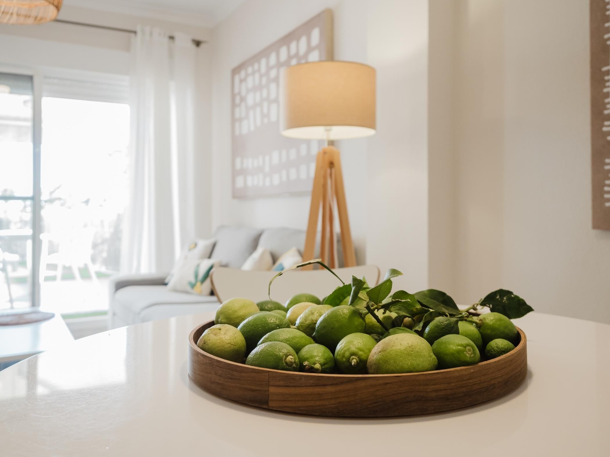 A wooden bowl of limes on a table in a bright room with a lamp and a sofa in the background.