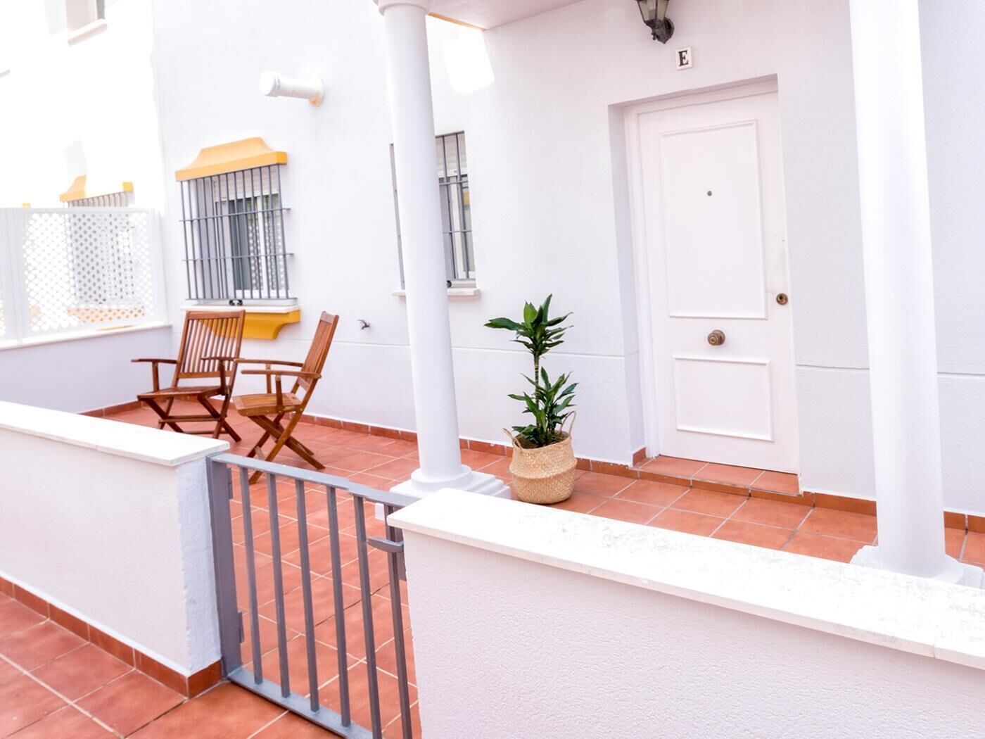 Un edificio blanco con una puerta marcada 'E', una ventana con reja de metal, una silla de madera, una planta en maceta y un patio con baldosas.
