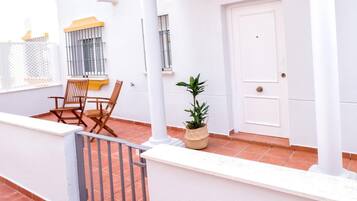 Un edificio blanco con una puerta marcada 'E', una ventana con reja de metal, una silla de madera, una planta en maceta y un patio con baldosas.