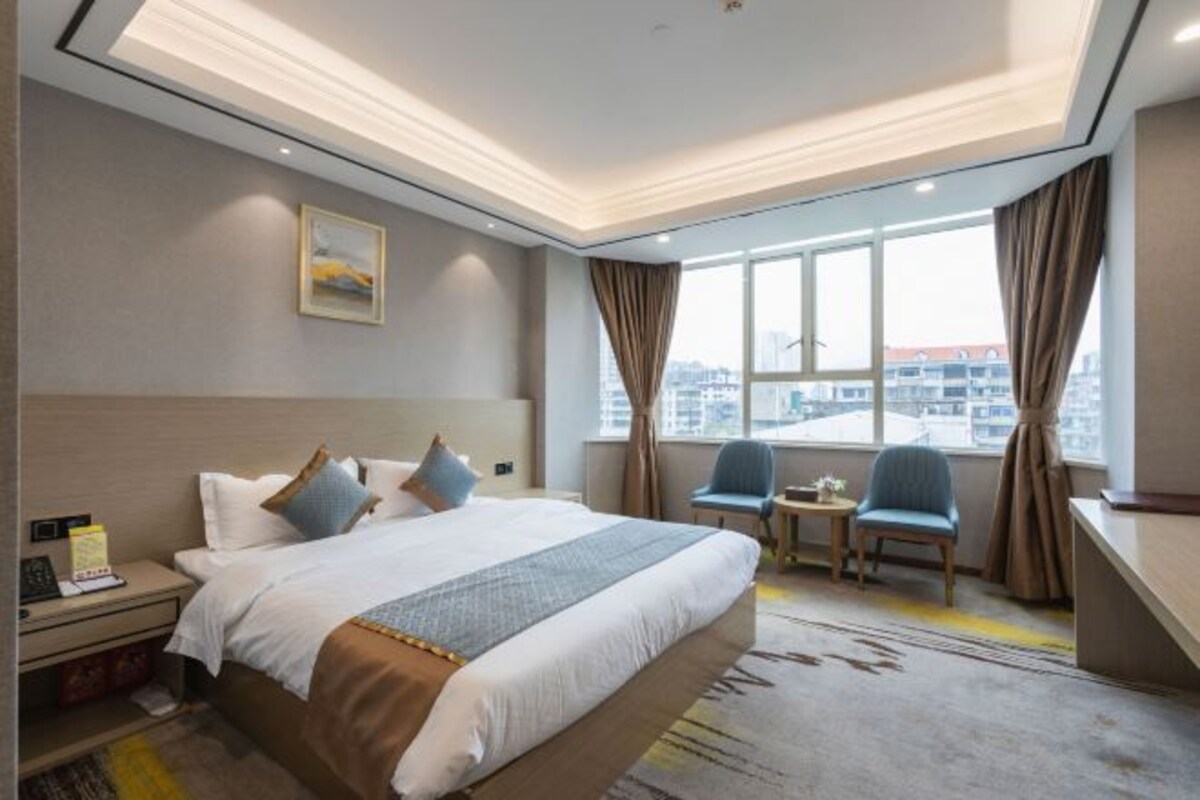 A hotel room with a large bed, a desk, two chairs, a small table, and a view of the city through large windows.