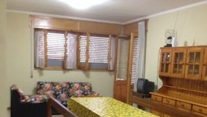 A dining area with a long table covered in a sunflower tablecloth, wooden benches, a floral sofa, and a television on a wooden cabinet. - Bosamarina House (Bosa)