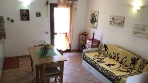 A living room with a sofa, a wooden dining table with a chair, a small side table with a vase, and a door with a curtain. - Appartamento Le Querce 4 (San Teodoro)