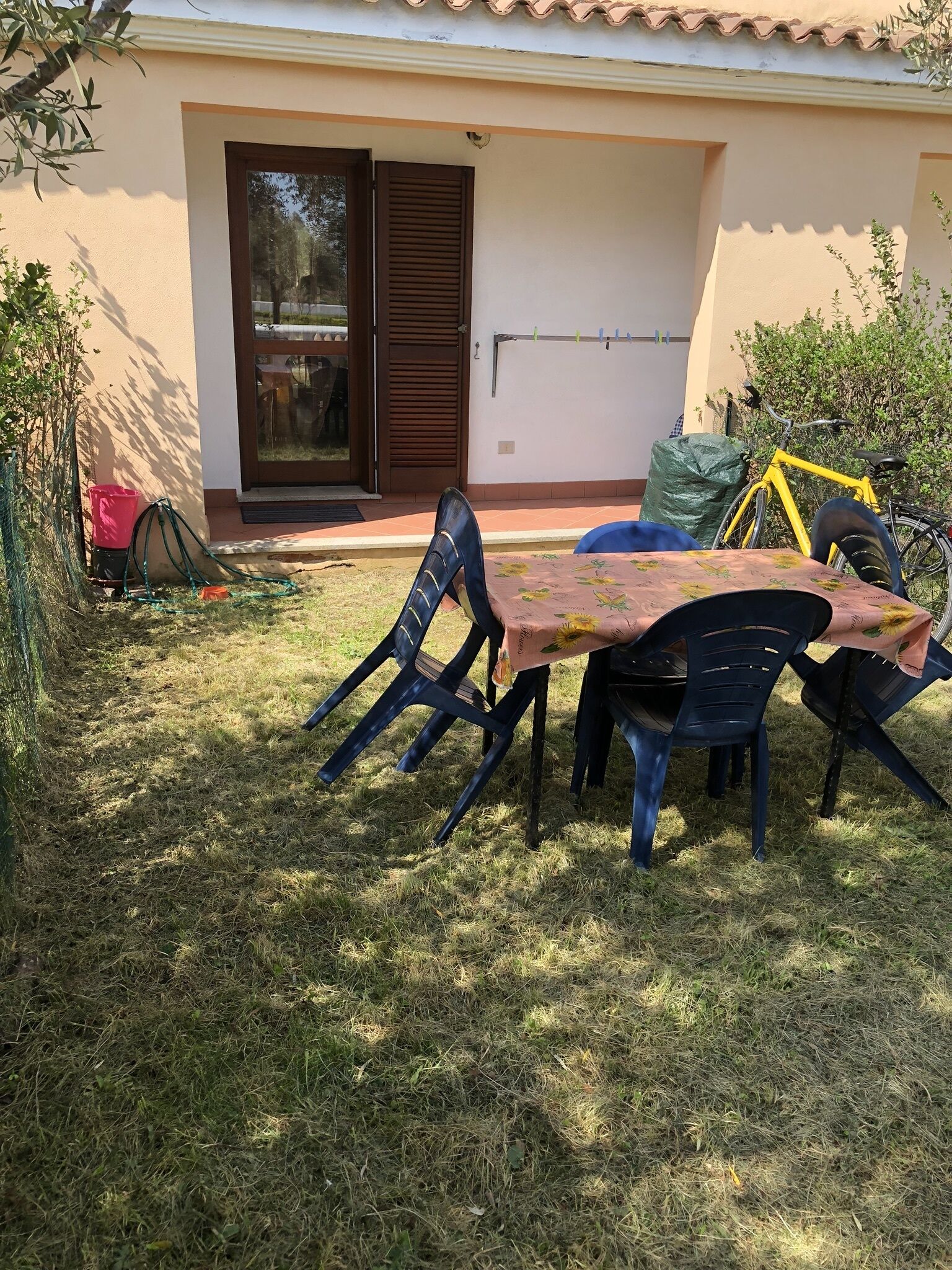 Un área de comedor al aire libre con una mesa y sillas, una bicicleta amarilla, y un edificio con tejado de baldosas y puerta corrediza.
