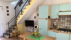 A dining area with a wooden table and chairs, a staircase with black railings, a kitchen with light blue cabinets, and a flat-screen TV on a stand. - Appartamento Le Querce 4 (San Teodoro)