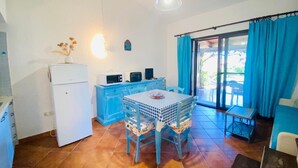 A compact kitchen with a refrigerator, microwave, and toaster, a blue table with a checkered tablecloth, and a sliding glass door leading to an outdoor area. - Appartamento Cala d'Ambra (San Teodoro)