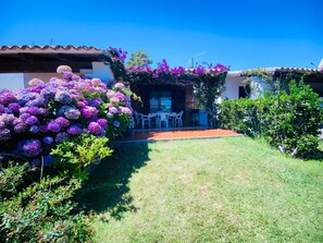 Un patio con mobiliario exterior, rodeado de exuberante follaje y plantas florecientes.