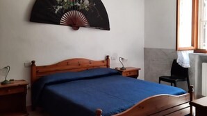 A bedroom with a wooden bed, a bedside table with a lamp, a wall-mounted fan, and a window with a view. - Magomadas Enjoy House (Magomadas)
