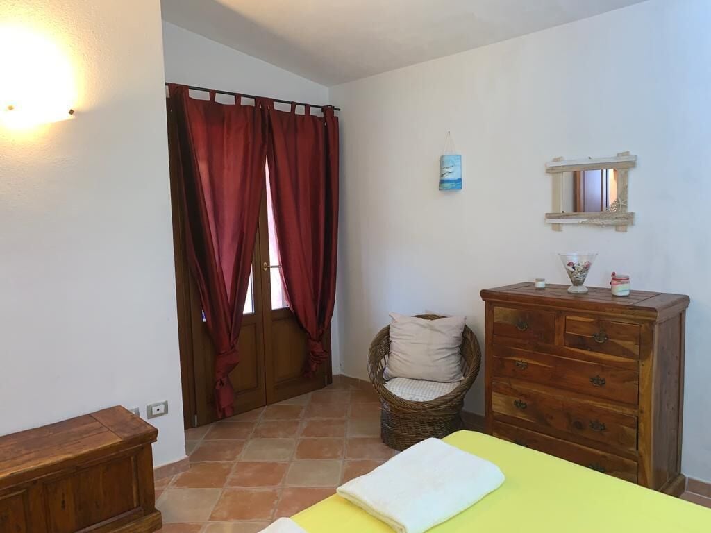 A bedroom with a bed, a wooden dresser, a wicker chair, and a window with red curtains.