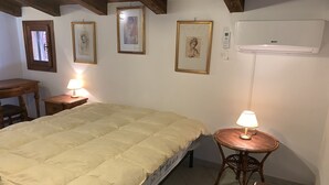 A bedroom with a bed, a desk, a chair, a round table with a lamp, and framed pictures on the wall.