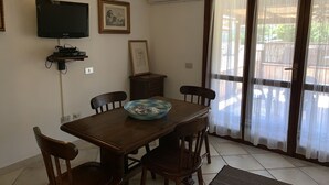 A dining area with a wooden table, four chairs, a television mounted on the wall, and a large window with curtains.