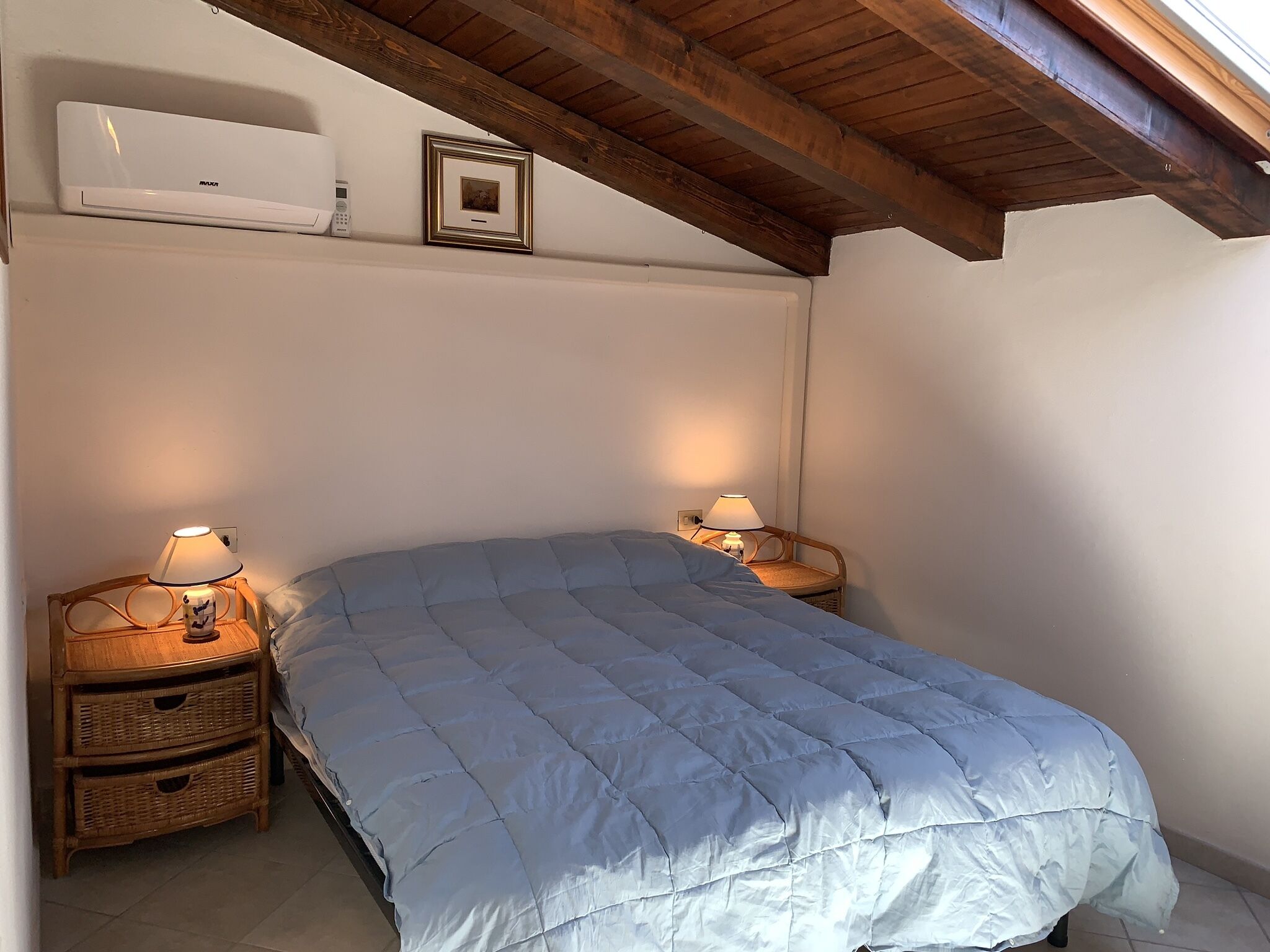 A bedroom with a bed, two bedside tables with lamps, a wall-mounted air conditioner, and a framed picture.