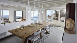 Un vaste espace dédié à la salle à manger, avec une longue table en bois, des chaises blanches, des luminaires suspendus et de grandes fenêtres offrant une vue sur la mer et les montagnes.