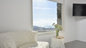Une chambre moderne avec un canapé blanc, une petite table d’appoint blanche ornée d’un vase, et une grande fenêtre offrant une vue sur des montagnes lointaines et l’eau.