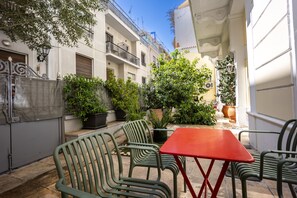 An outdoor patio with green chairs and a red table, surrounded by potted plants and a white building.