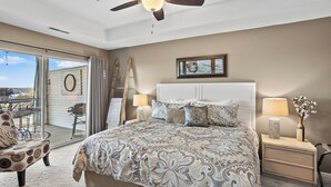 Une chambre à coucher comprenant un lit, deux tables de chevet, un ventilateur de plafond, une grande fenêtre avec vue et un fauteuil.