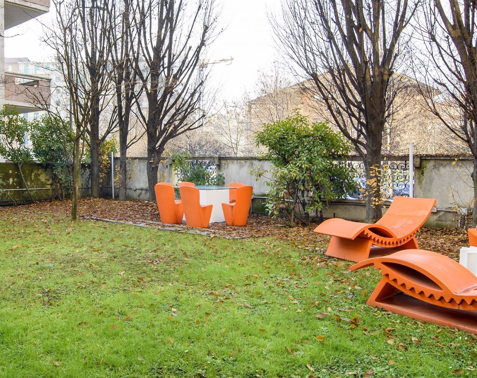 Un espacio al aire libre con césped, una mesa y sillas, rodeado de árboles y un muro bajo.