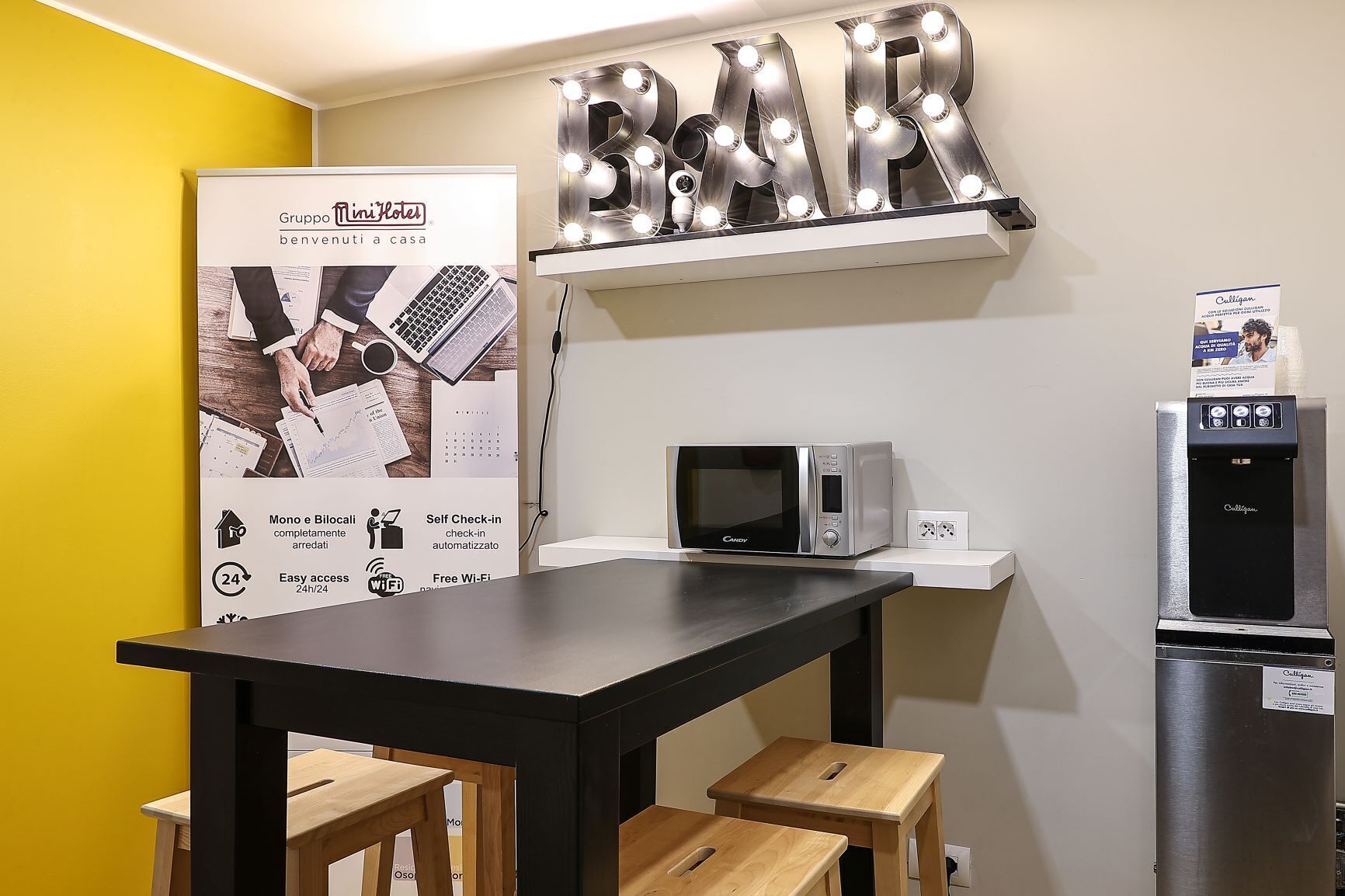 A kitchenette with a microwave, a water dispenser, and a refrigerator. There is a 'BAR' sign above the counter and a promotional poster on the wall.