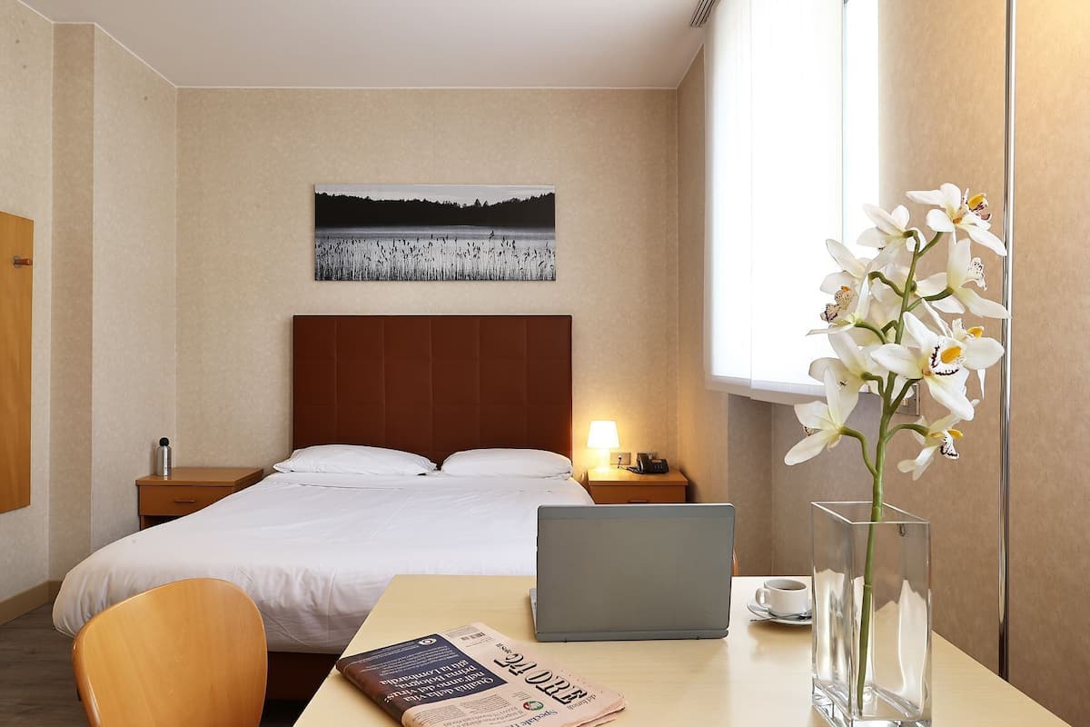 A hotel room with a bed, a desk with a laptop, a vase with flowers, and a painting on the wall.