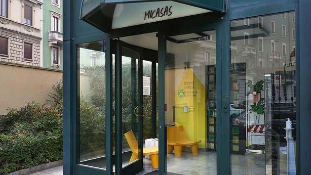 A modern building entrance with a glass door and a yellow bench outside. - Residence Cimarosa (Milan)