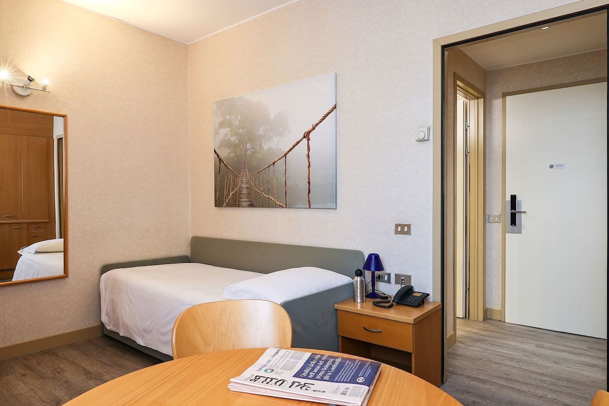 A hotel room with a bed, a desk with a lamp, a chair, a table with a newspaper, a painting of a bridge, and a door.