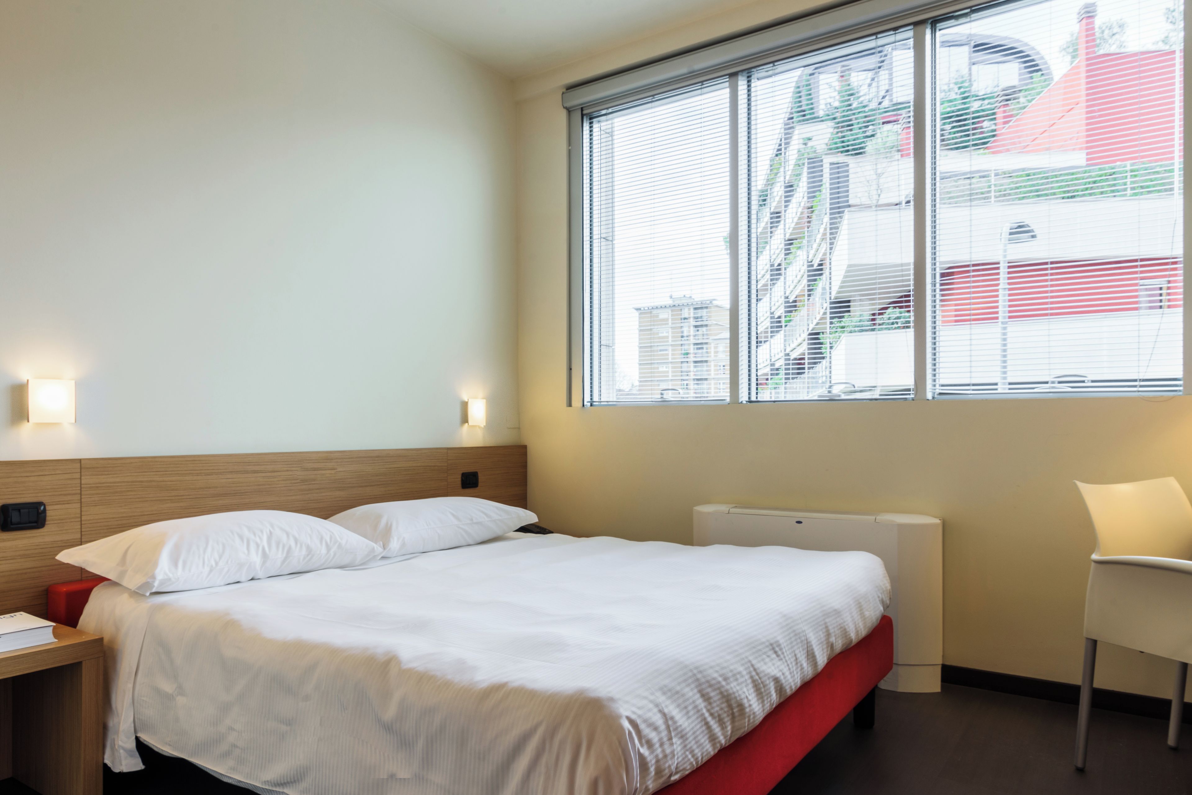 A hotel room with a bed, a nightstand, a chair, a window with blinds, and a radiator.