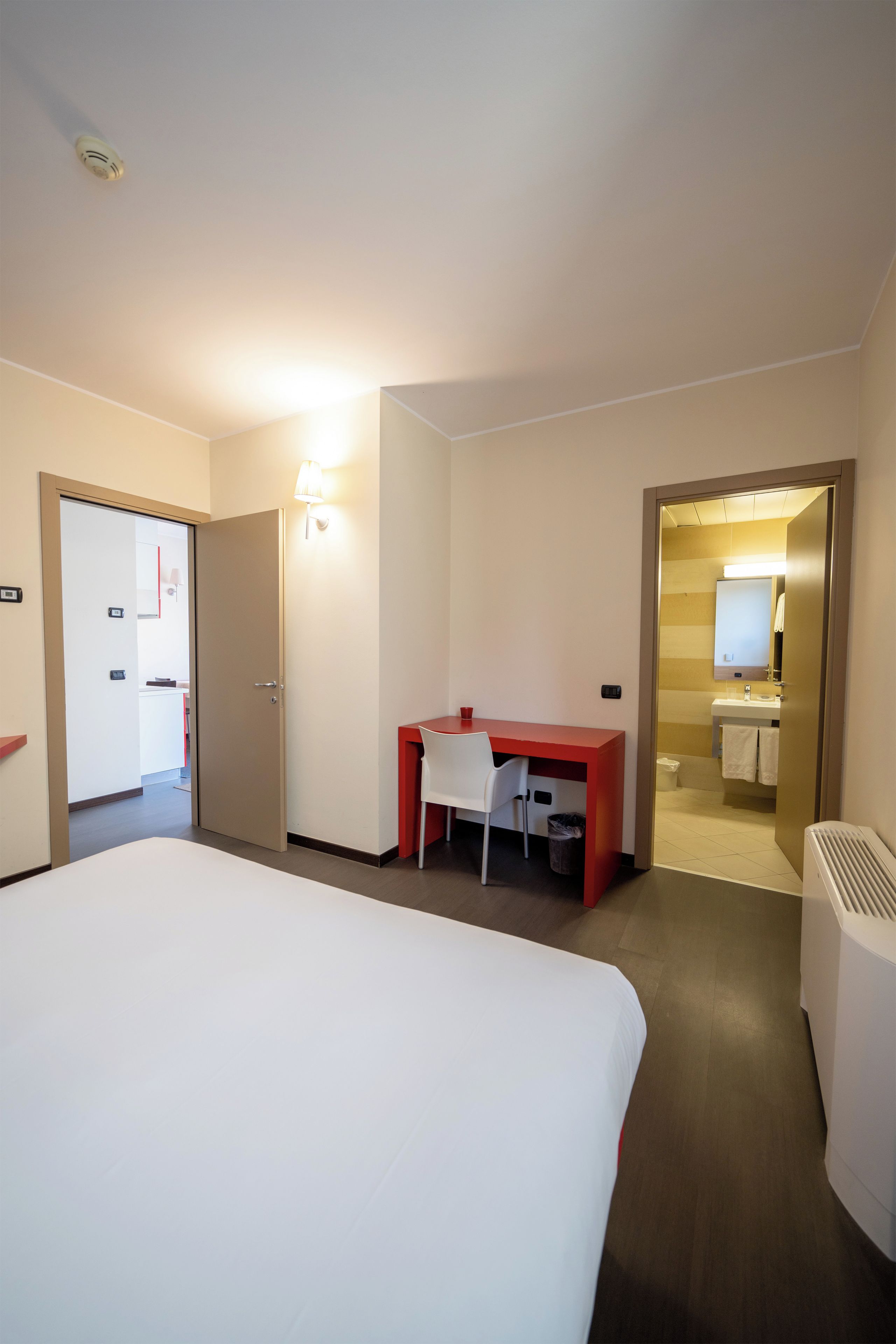 Ein Hotelzimmer mit einem großen Bett, einem Schreibtisch mit Stuhl, einem Badezimmer mit Spiegel und einer Tür zu einem weiteren Raum.