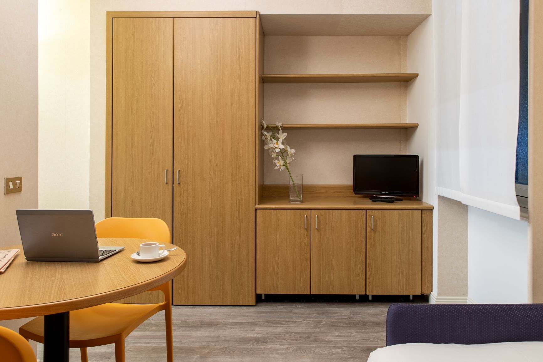 A hotel room with a wooden wardrobe, a desk with a laptop, a chair, a small table with cups, a TV on a stand, and a bed.