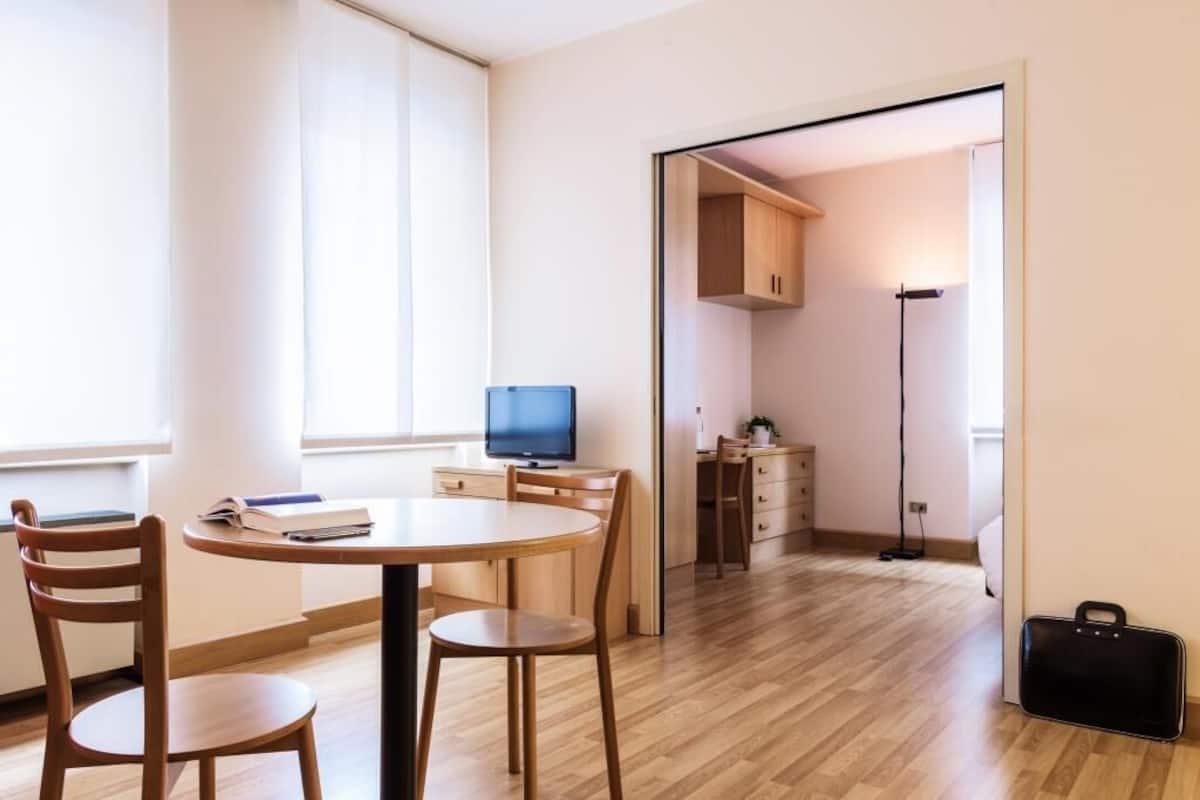 A modern, well-lit room with a round wooden table, two chairs, a television, and a kitchen area visible through an open door.