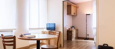 Una habitación moderna y bien iluminada con una mesa redonda de madera, dos sillas, un televisor y una cocina visible a través de una puerta abierta.