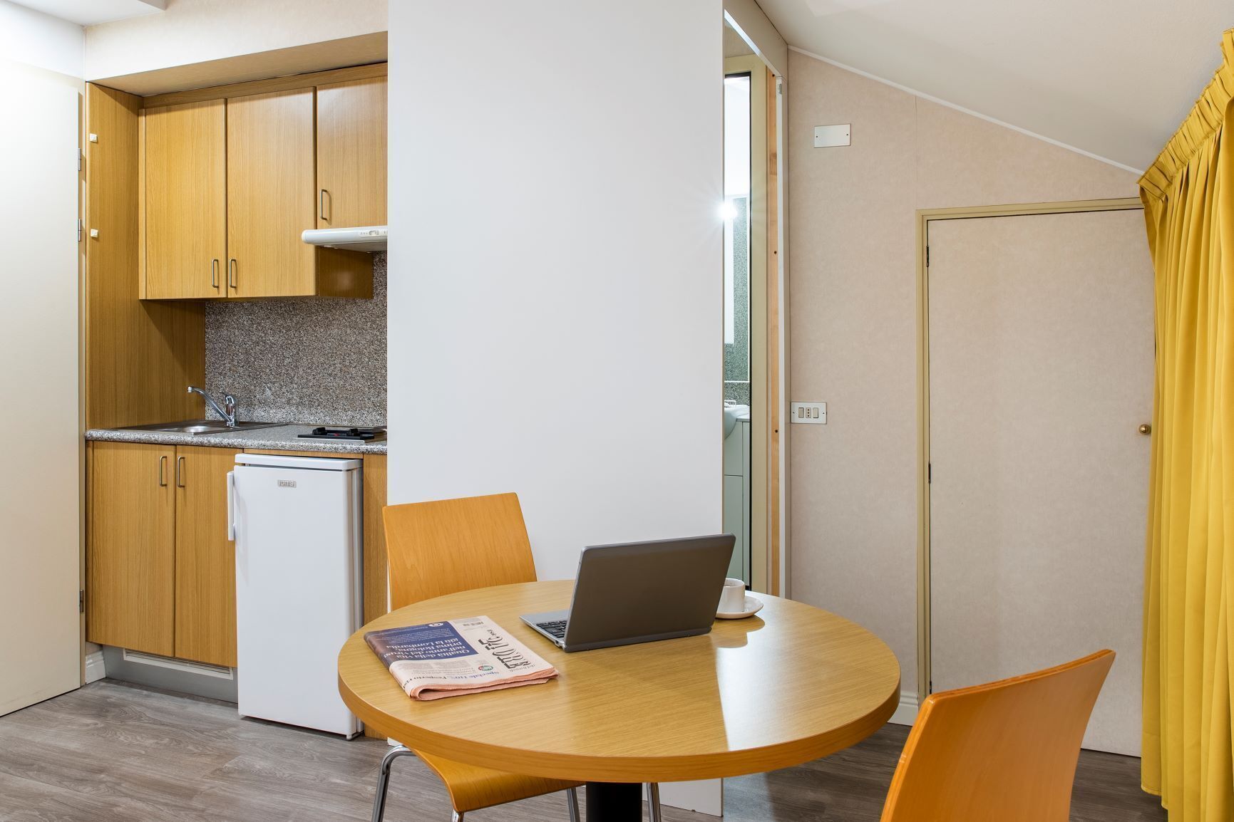 Una cocina compacta con armarios de madera, un refrigerador blanco y una pequeña mesa de comedor con una computadora portátil y un periódico.