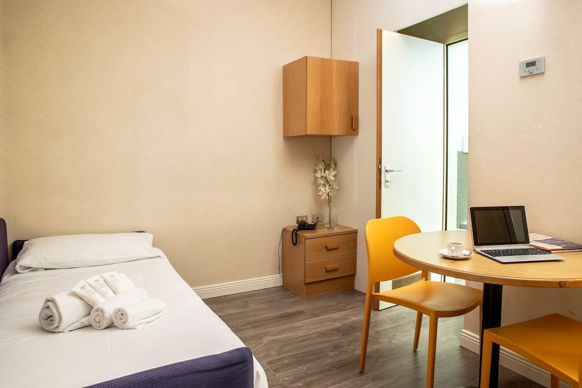 A hotel room with a bed, a desk with a laptop, a chair, a nightstand, and a door leading to a balcony.