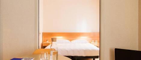 A hotel room with a bed, a desk with a laptop, a glass of water, and a television.
