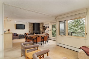 A spacious living room with a fireplace, a dining area, and large windows offering a view of trees.