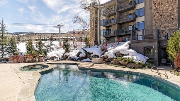 Zona de piscina con agua cristalina, rodeada por un edificio de piedra con varios balcones, y una vista de paisaje nevado con un telesilla al fondo.