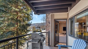 Un balcón con una parrilla, una silla Adirondack azul y vistas a árboles y edificios.