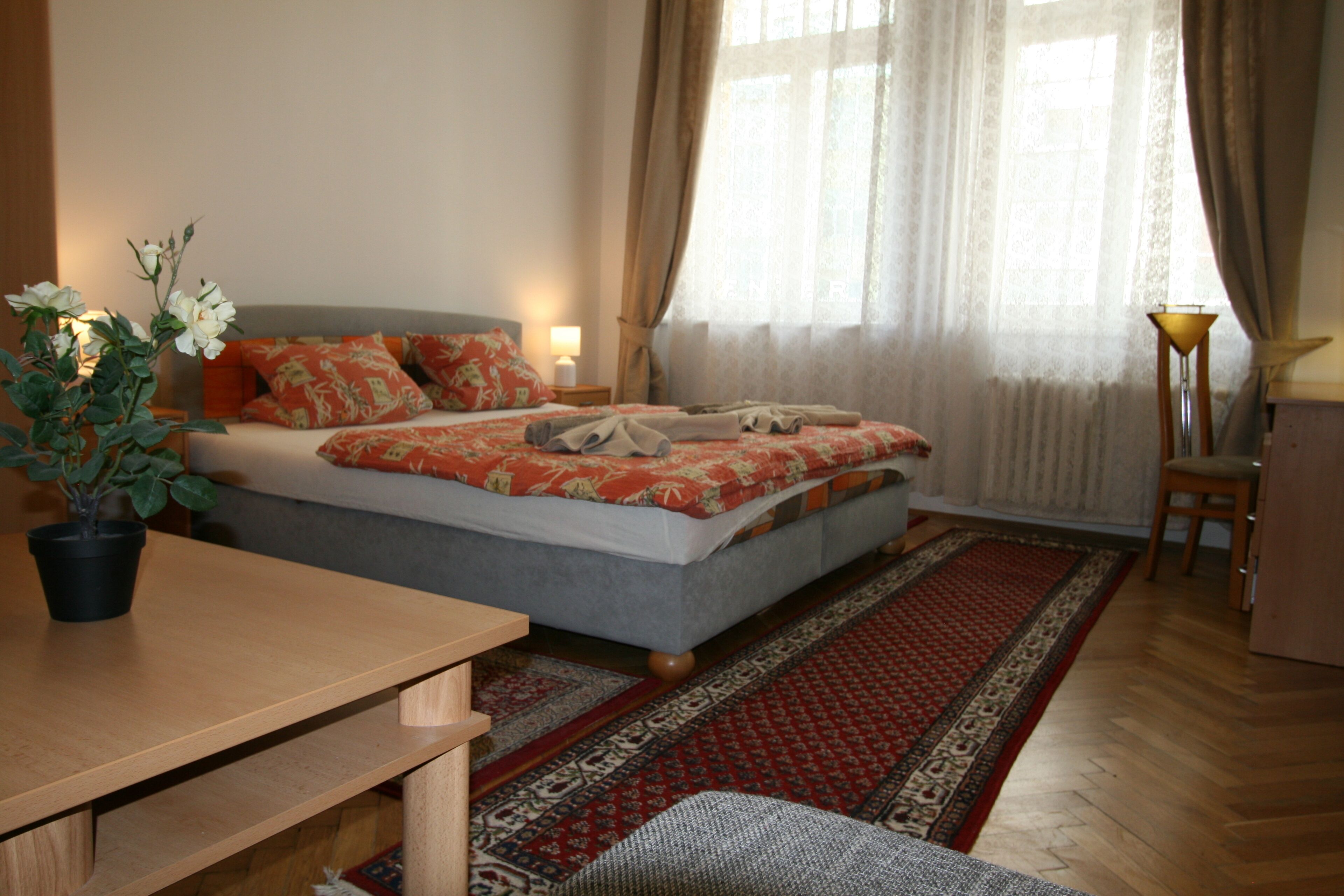 A bedroom with a bed, a bedside table, a lamp, a chair, a potted plant, and a window with curtains.