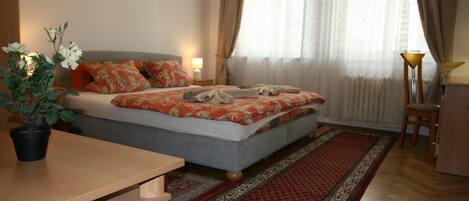 A bedroom with a bed, a bedside table, a lamp, a chair, a potted plant, and a window with curtains.