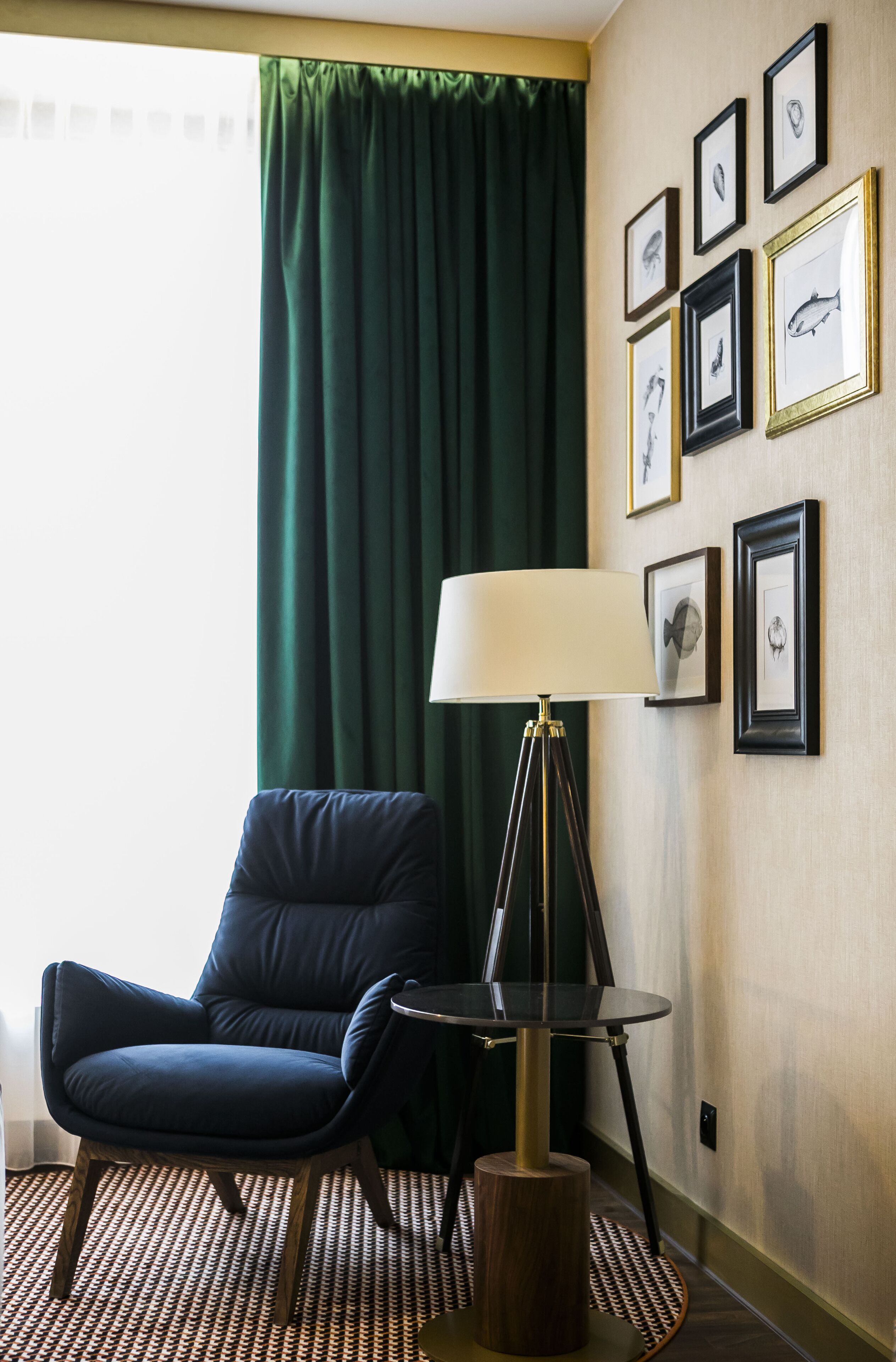 Una habitación con un sillón azul oscuro, una mesita redonda con una lámpara y una alfombra estampada. En la pared hay cuadros enmarcados.