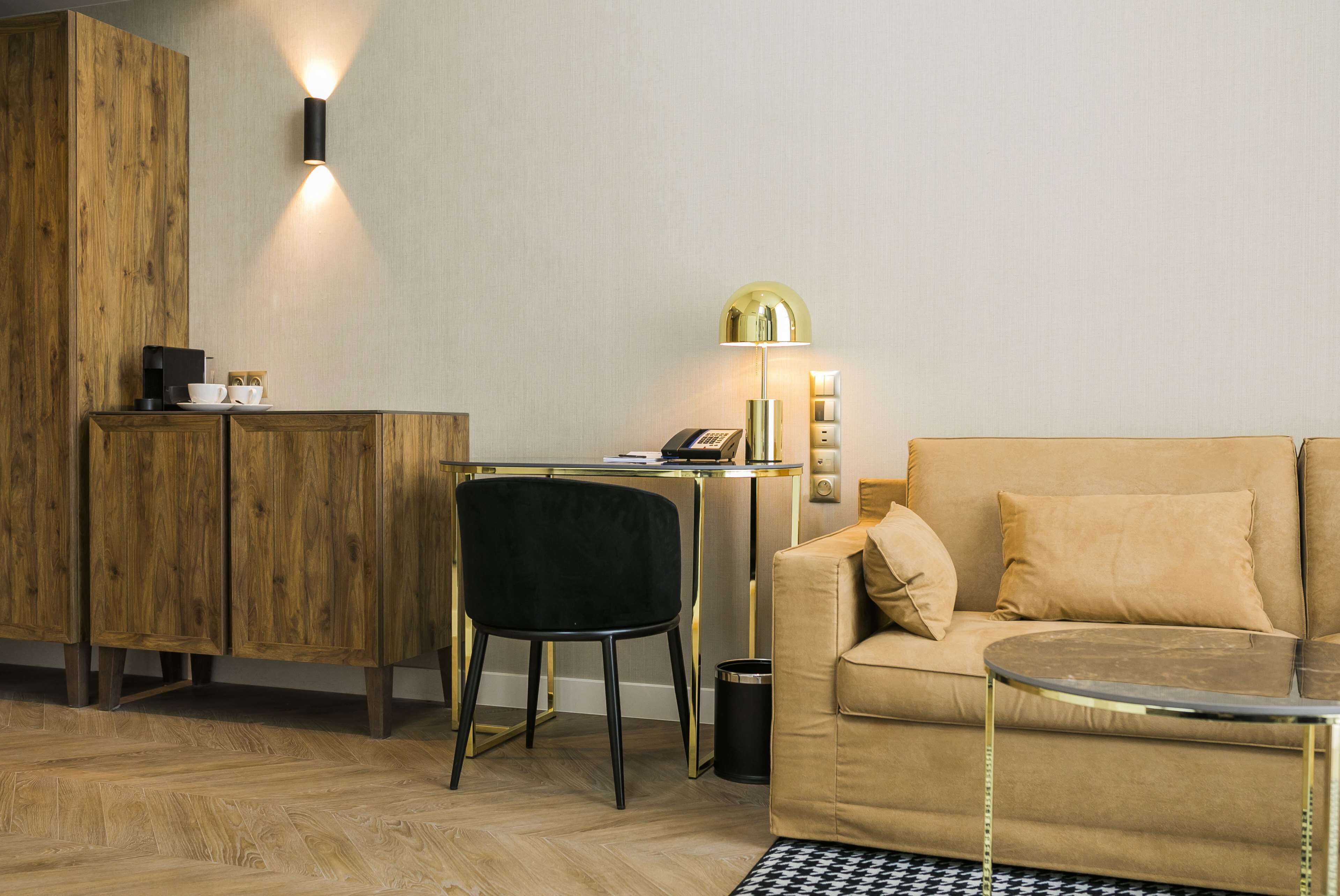 Una sala de estar moderna con un aparador de madera, una silla negra, un sofá color beige, una mesita redonda pequeña y un escritorio con una lámpara.