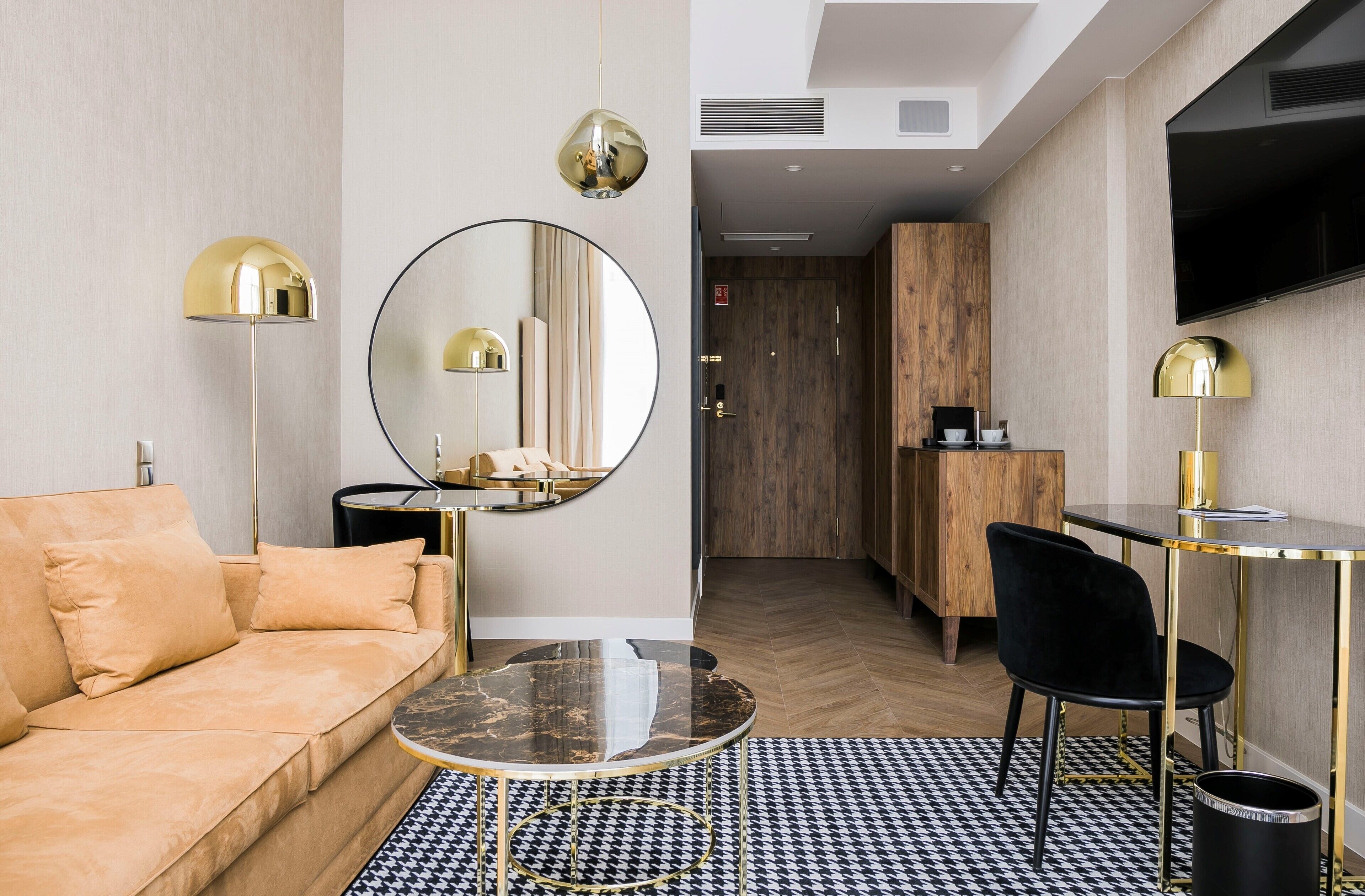 Habitación de hotel moderna con sofá, espejo redondo, mesa de comedor y televisor montado en la pared.