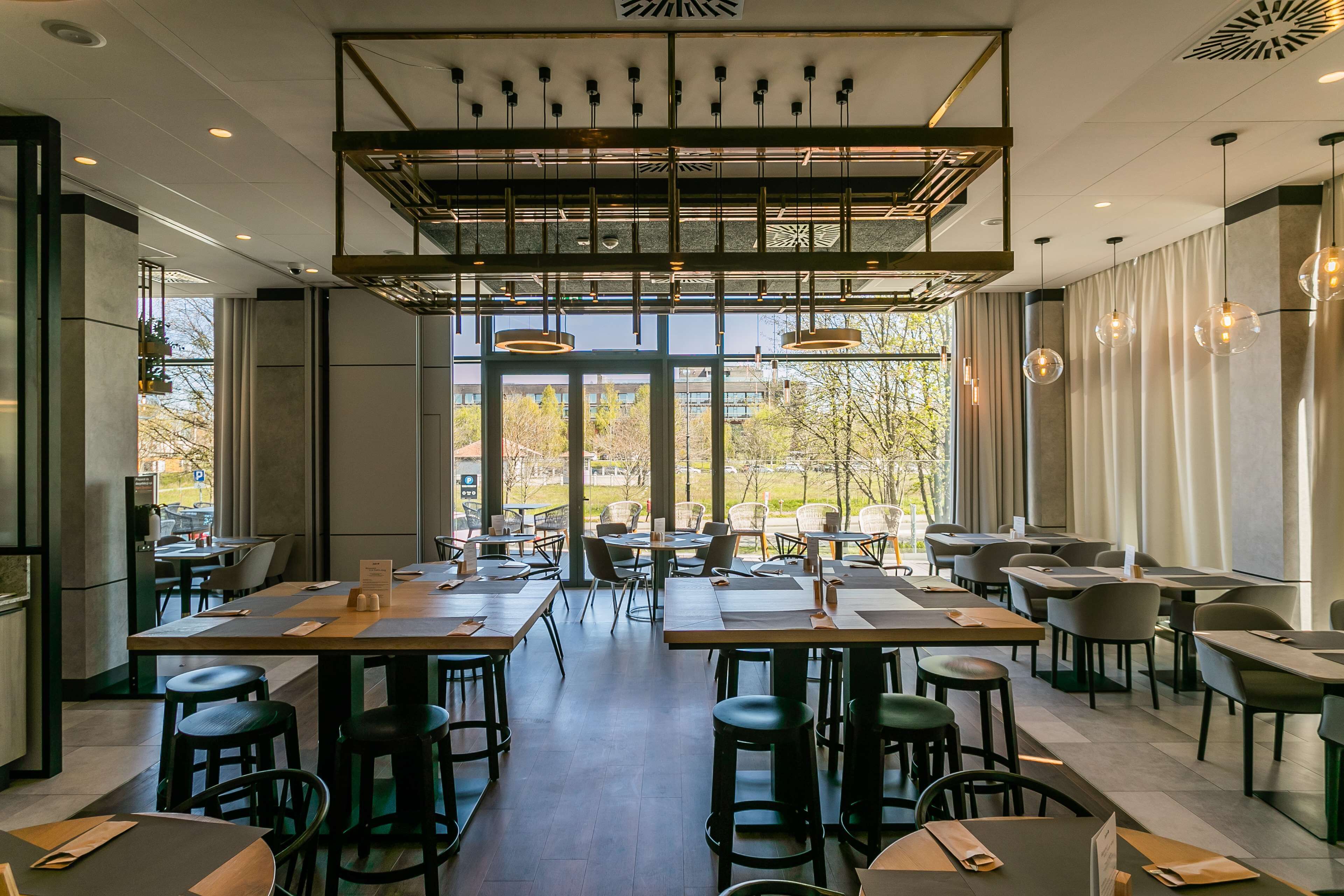 A modern restaurant with high ceilings, pendant lighting, and large windows offering a view of the outdoors.