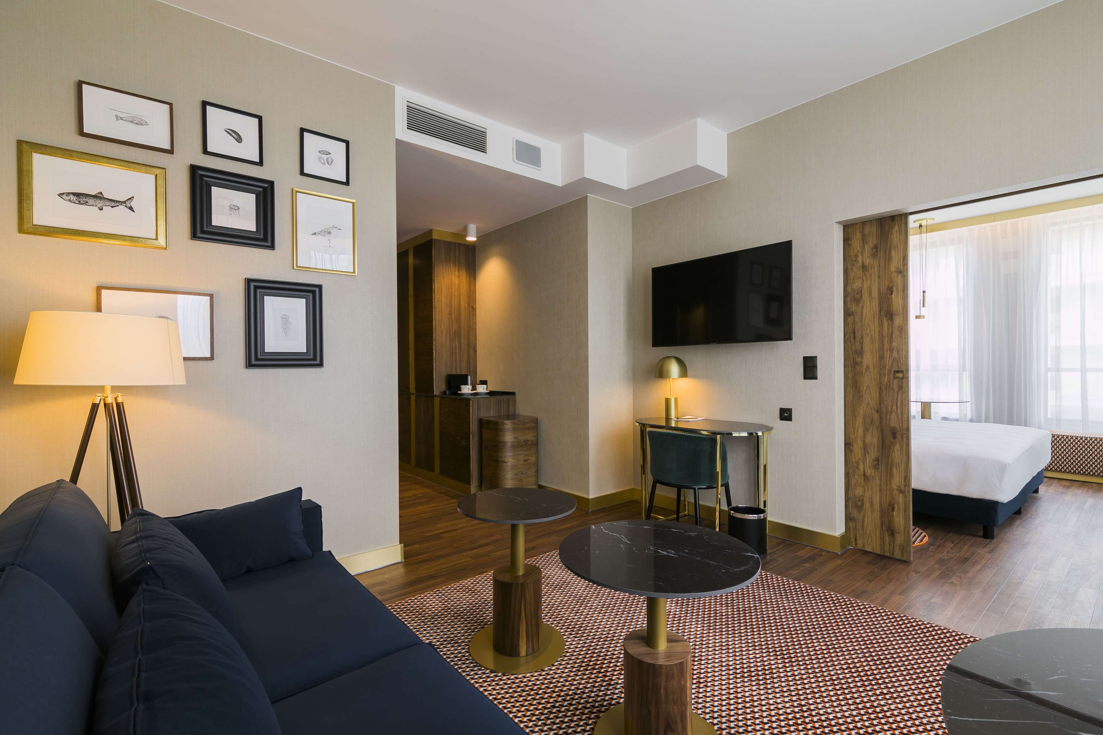 Une chambre d’hôtel moderne avec un canapé, une table ronde, un téléviseur et des tableaux encadrés au mur.