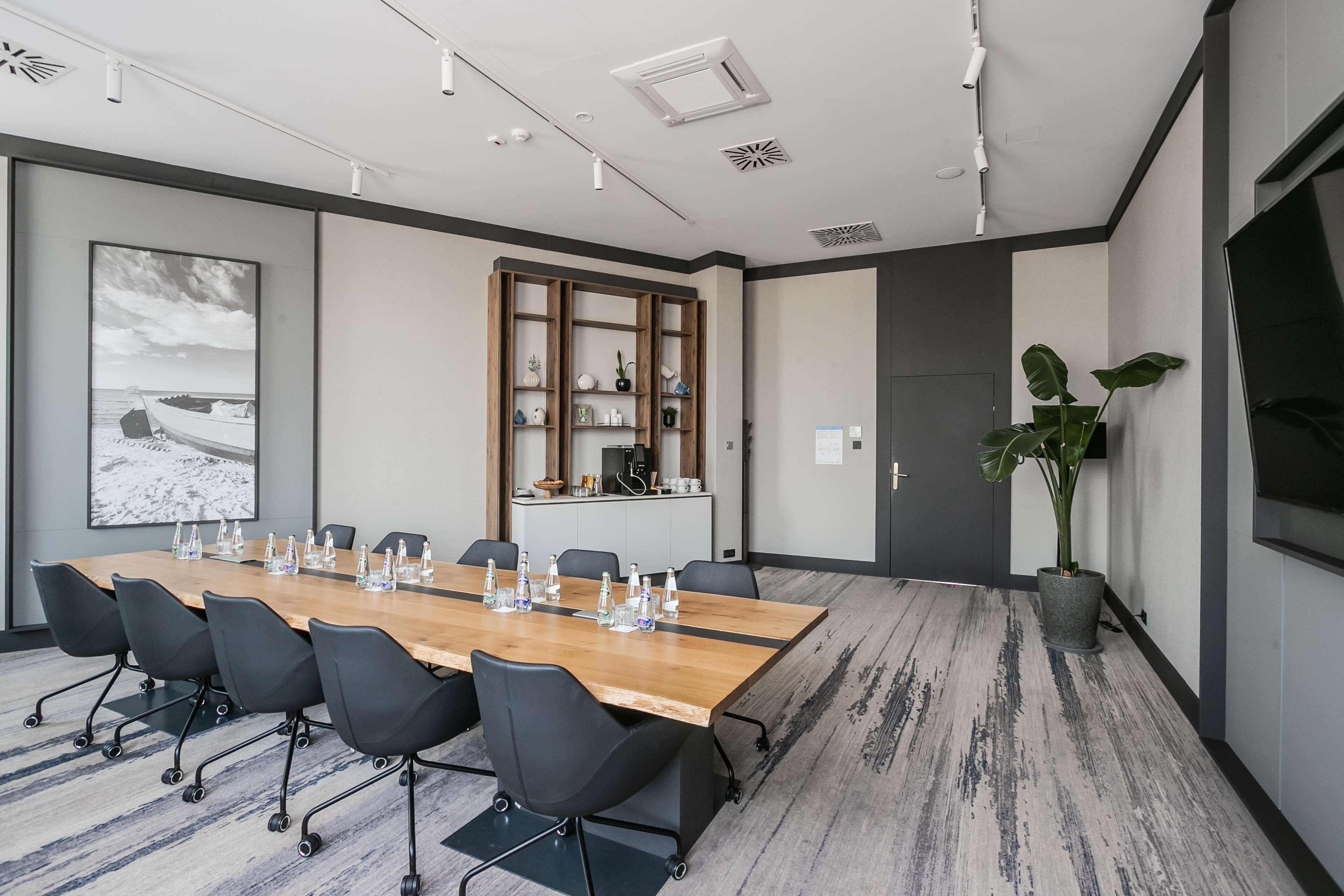 Een vergaderruimte met een lange tafel, zwarte stoelen, een houten kast en een potplant.